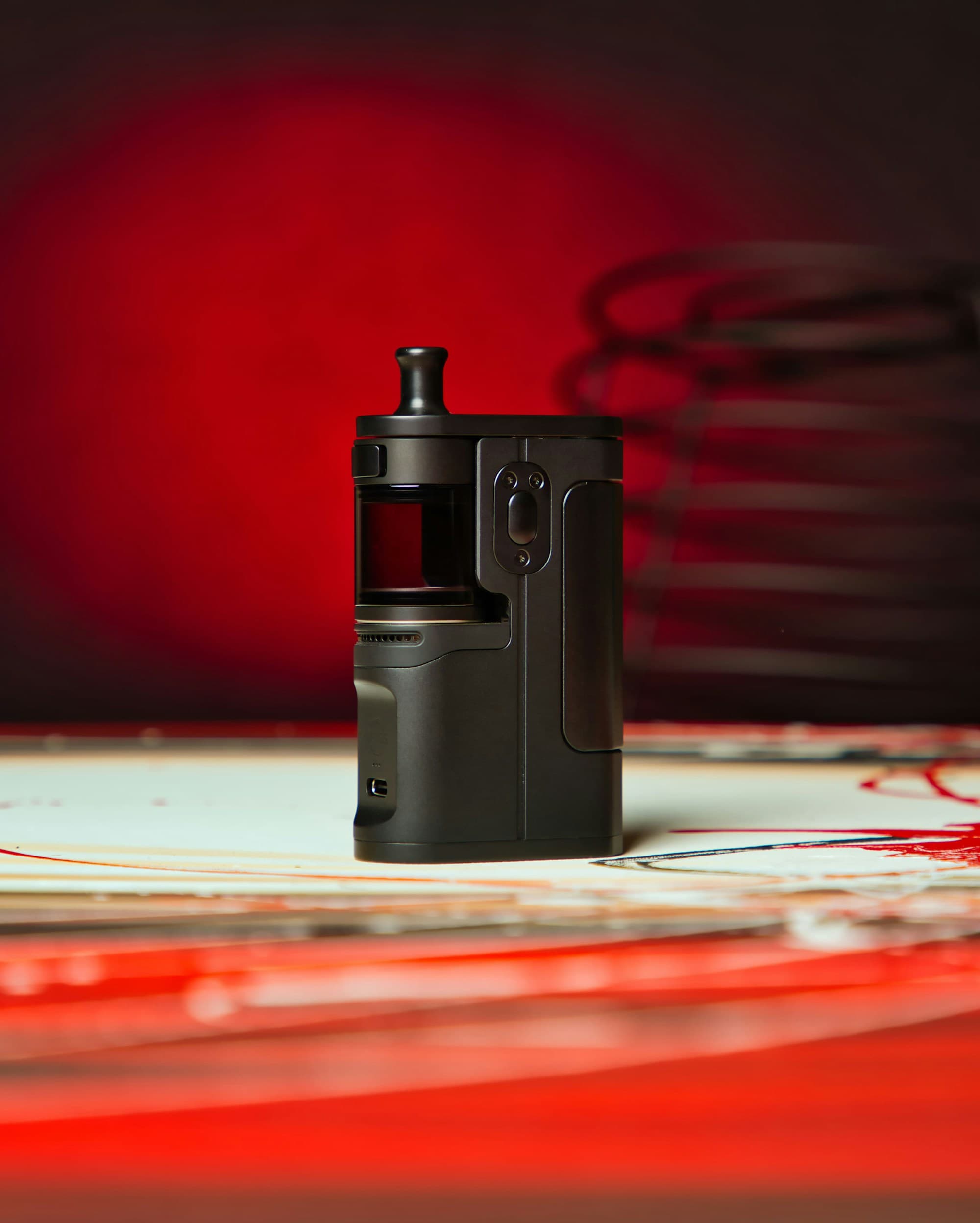 A matte black rechargeable pod vape device standing upright against a deep red and black studio backdrop